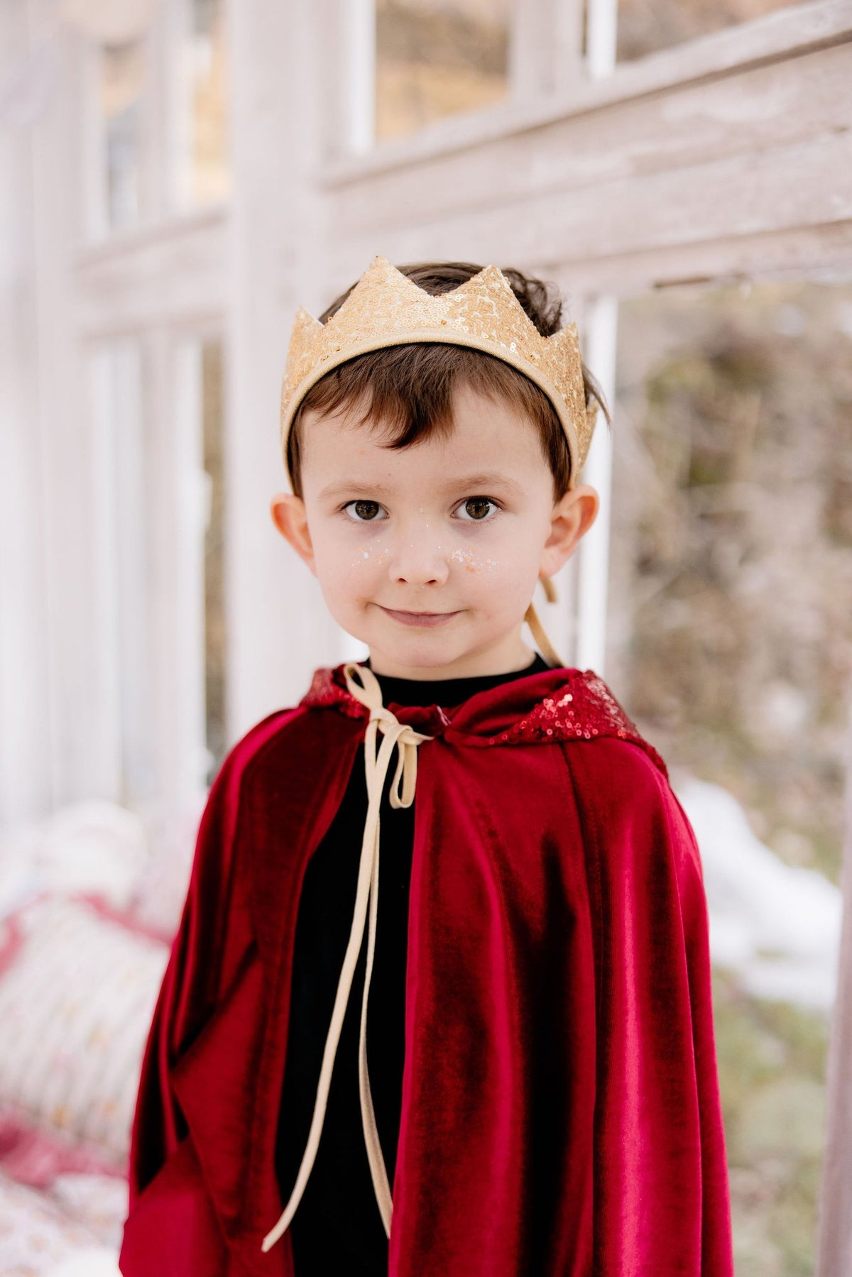 5. Boy wearing a red velour cape and gold crown, standing indoors with a soft background