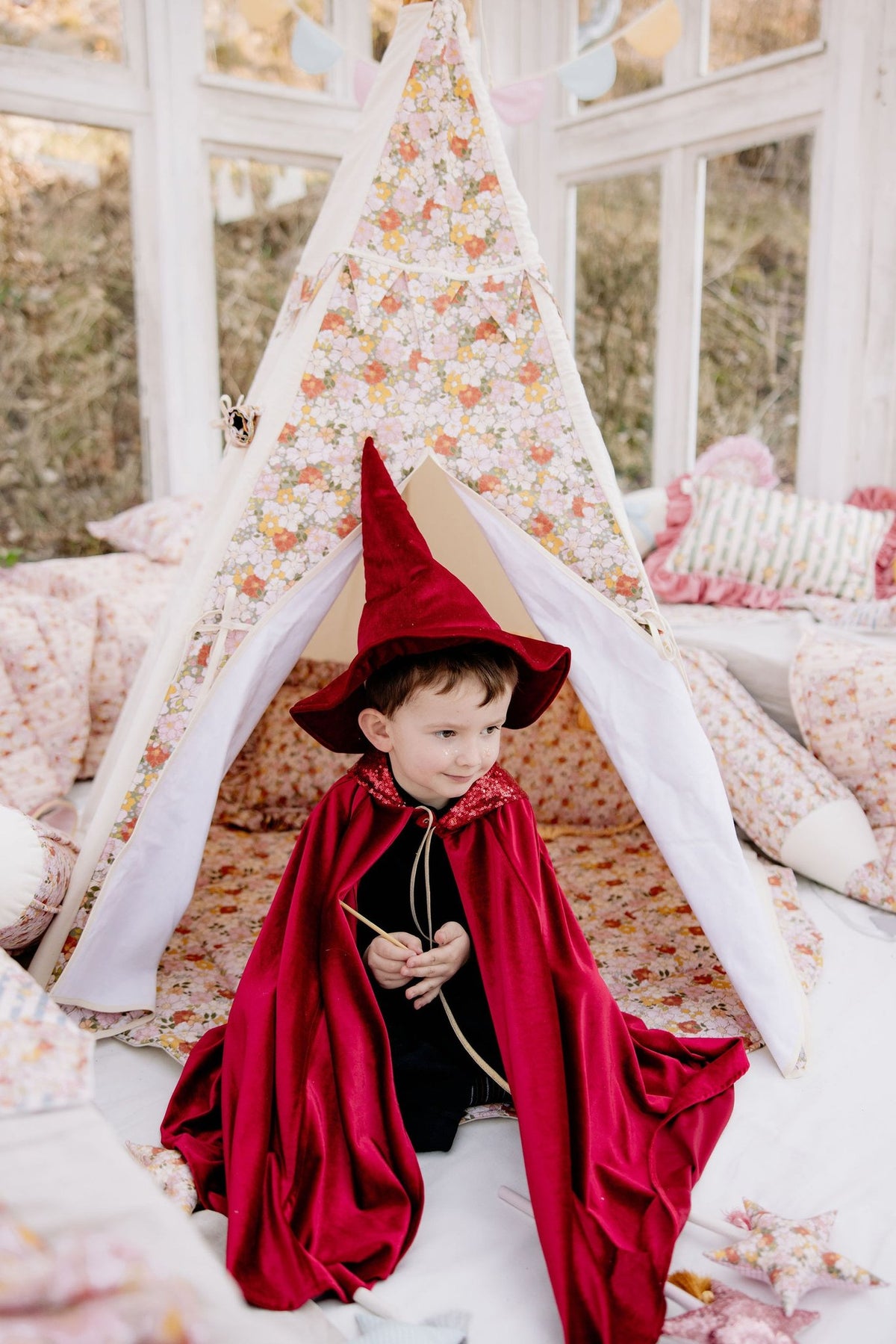 9. Boy in red velour cape and wizard hat sitting in front of a floral tent indoors