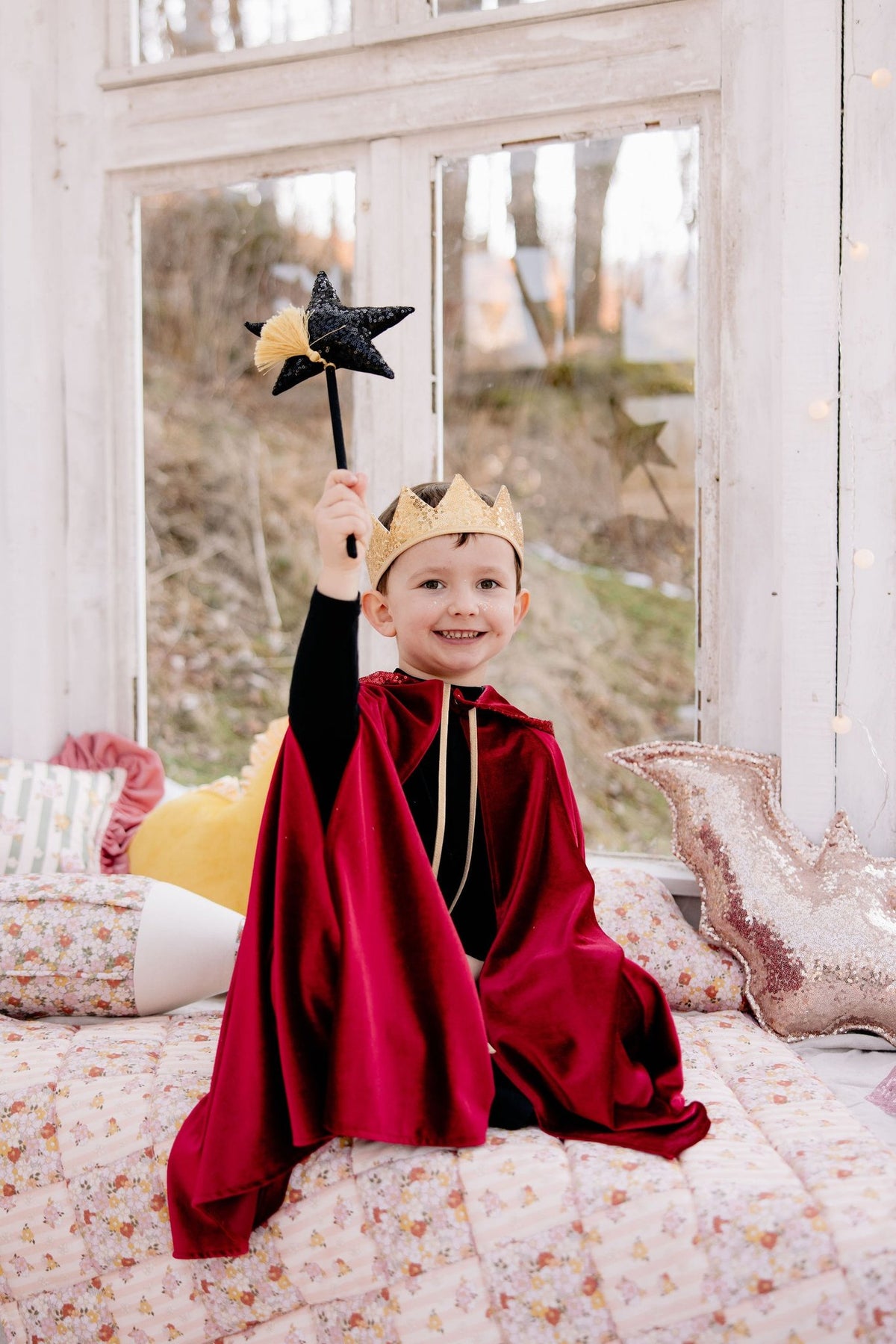 2. Boy in red velour cape and gold crown holding a star wand, sitting on a bed in a cozy room with large windows