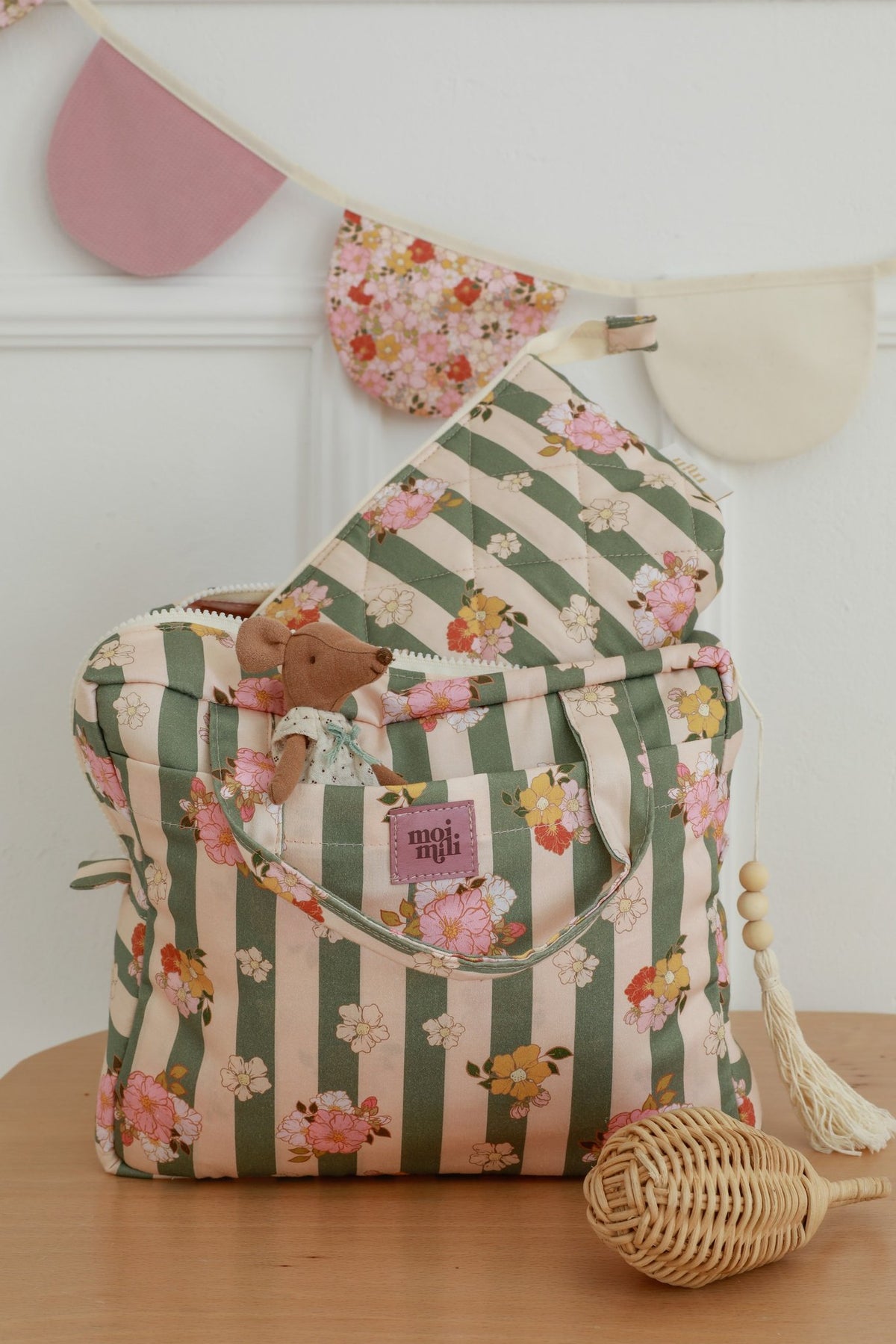 4. Green striped floral makeup bags with Moi Mili logo on wooden table, one bag inside the other, with decorative bunting