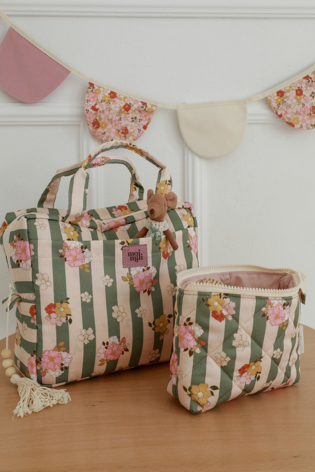 1. Two green striped makeup bags with floral print on wooden table, featuring Moi Mili logo, against a decorative wall with bunting