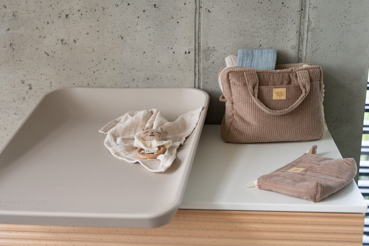 4. Latte-colored makeup bag set of two on a white surface next to a changing table, featuring large and small bags with Moi Mili logo.