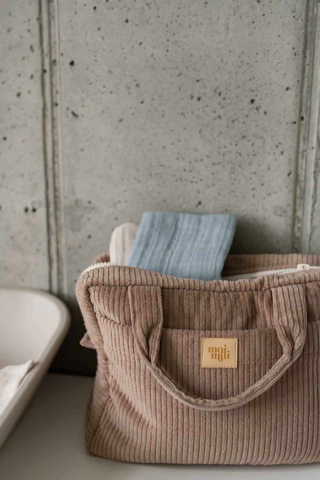 3. Large latte-colored makeup bag with Moi Mili logo on a shelf, partially filled with items, against a concrete wall.
