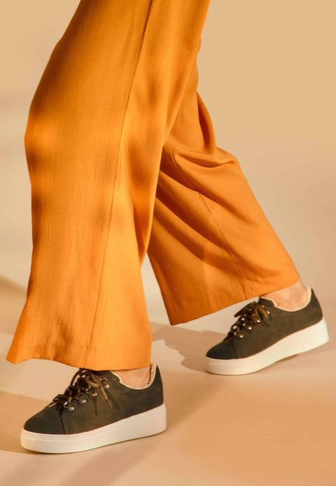 2. Close-up of Mangará Araucaria Warm Sneakers in green, worn by a woman with orange pants, showcasing suede material and platform heel