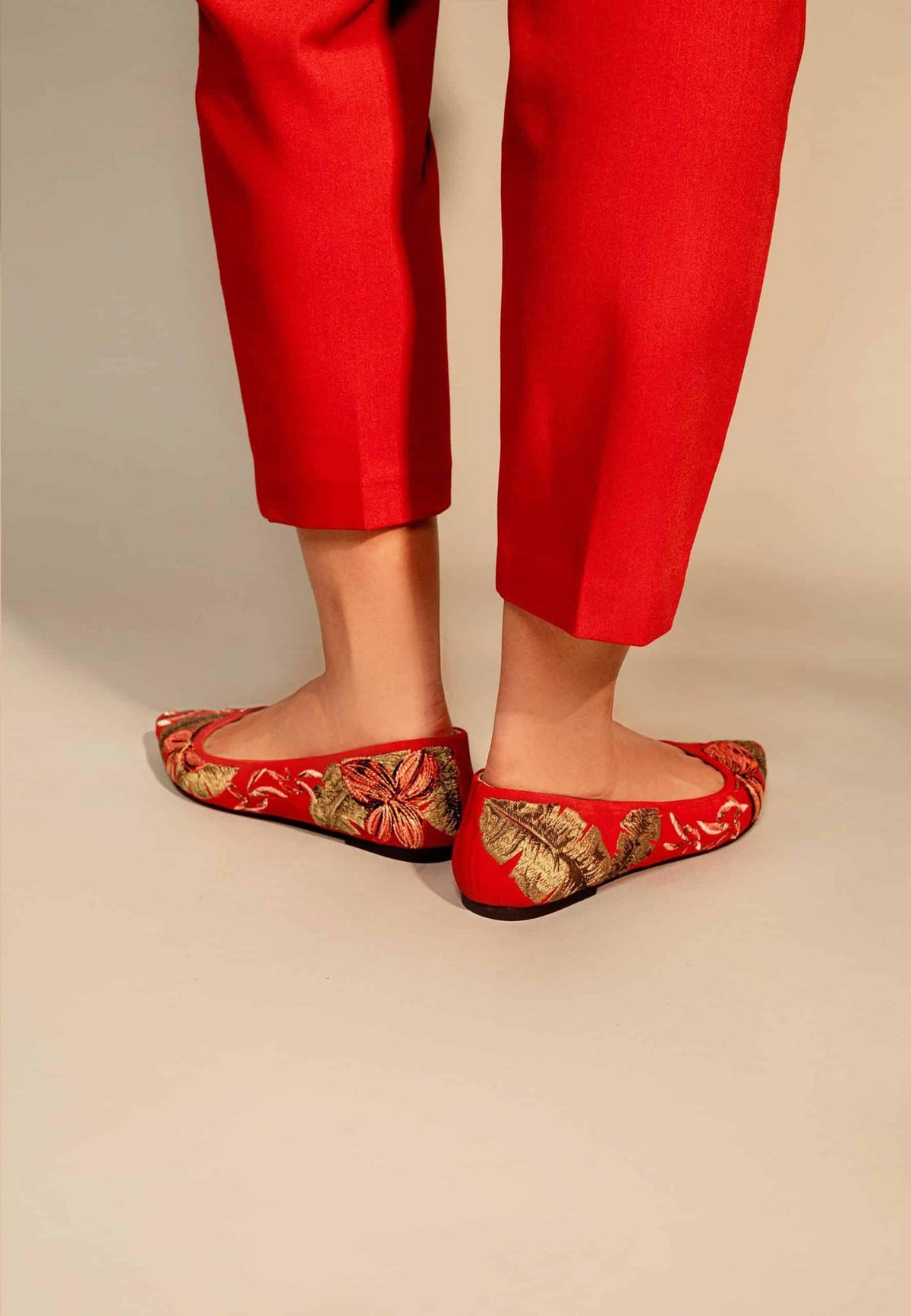 2. Woman sitting wearing Mangará Pindaiva Ballerinas in red suede with floral embroidery, paired with red pants and white shirt