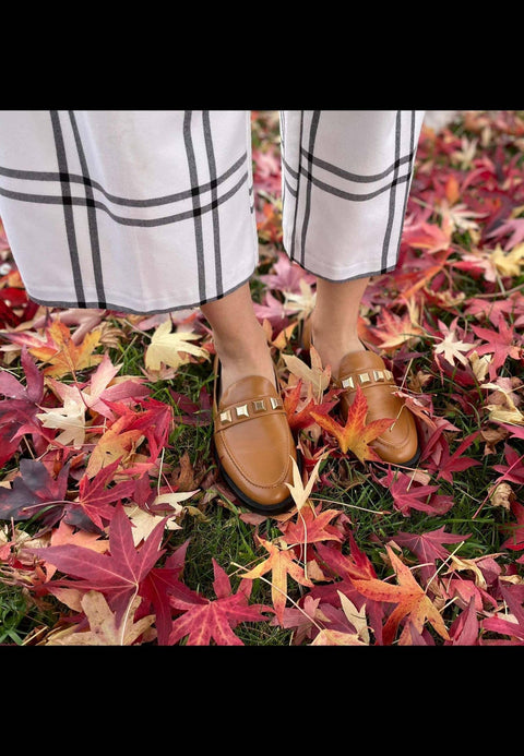 1. Women's Mangará Caete leather loafers in brown with metal studs, worn by a model standing on autumn leaves