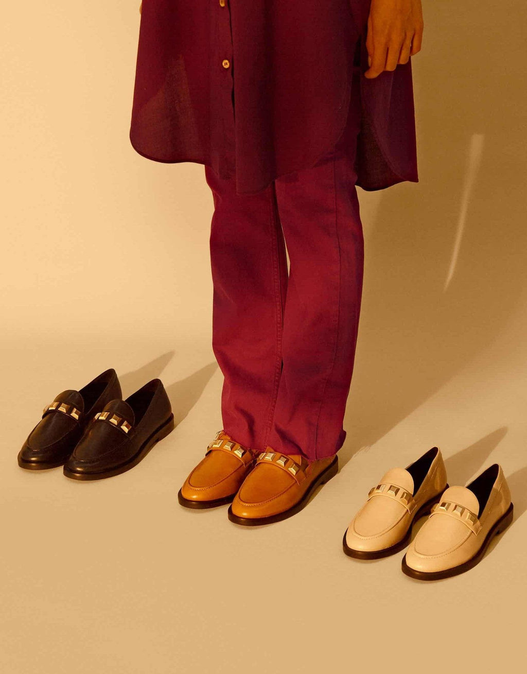 1. Women's Mangará Caete leather loafers in black, brown, and beige with metal studs, worn by a model in purple pants, studio setting