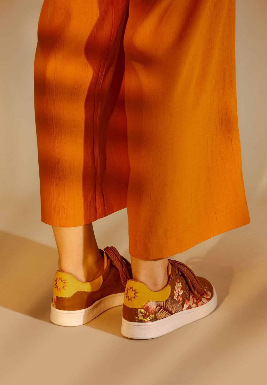1. Women's Mangará embroidery suede sneakers in brown, worn with orange pants, shown from the back in a lifestyle setting