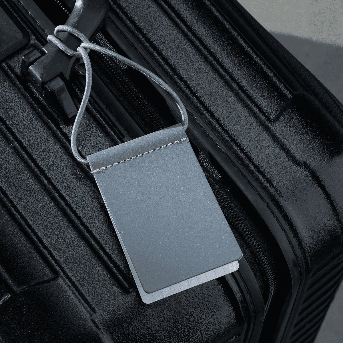 2. Grey MARCH Reflective Luggage Tag attached to black suitcase, highlighting durability
