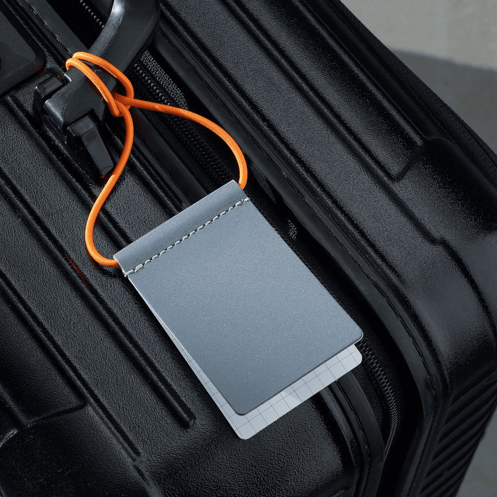 7. Grey MARCH Reflective Luggage Tag with neon orange strap on black suitcase, highlighting attachment