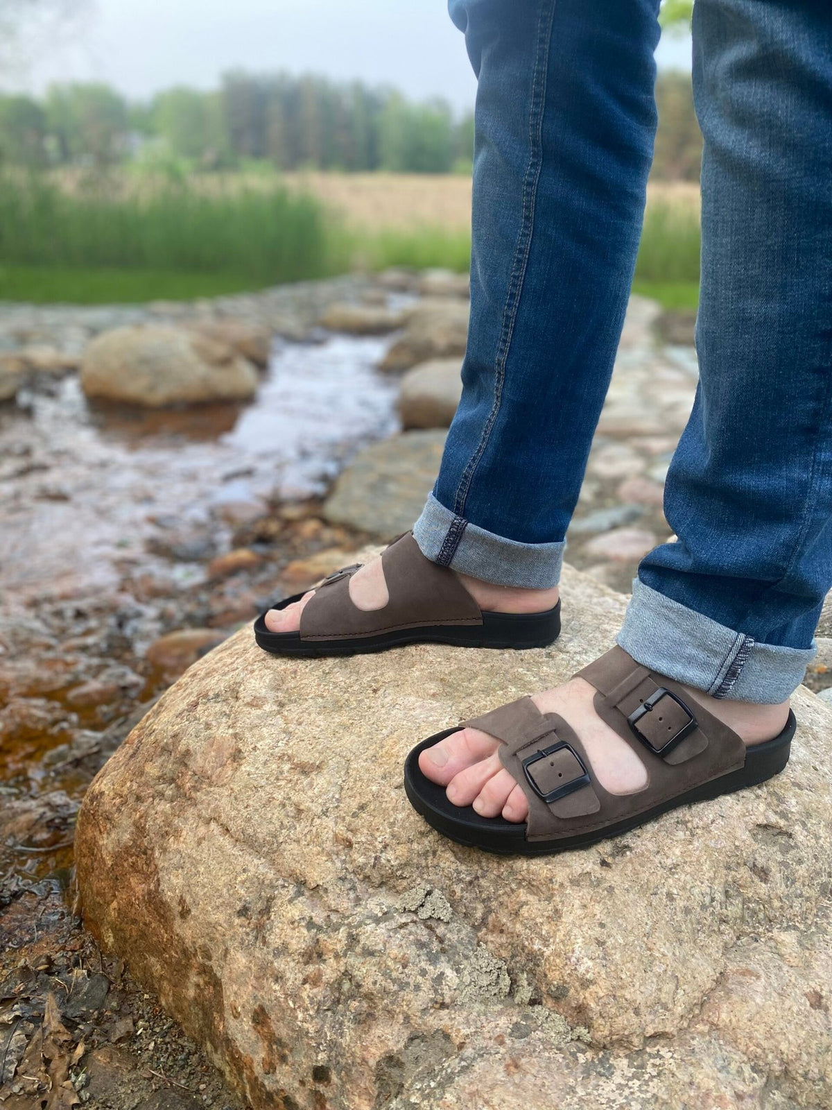1. Men's dark brown leather sandals by Omaking worn by a model standing on a rock by a stream