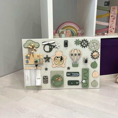 1. Montessori busy board in natural setting with interactive elements like gears, locks, and zippers, designed for sensory play and skill practice, featuring green and beige tones, by BusyBunnyToys.
