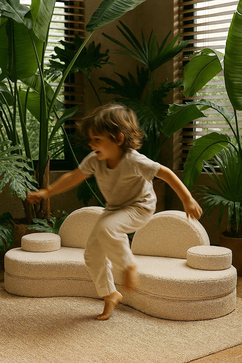 1. Child jumping on modular kids' sofa in Bearly off-white, surrounded by lush green plants in a cozy room