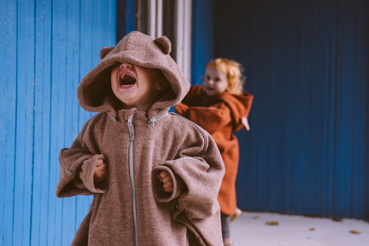 1. Two children wearing Patulove merino wool car ponchos with bear ears, playing outdoors against a blue wall