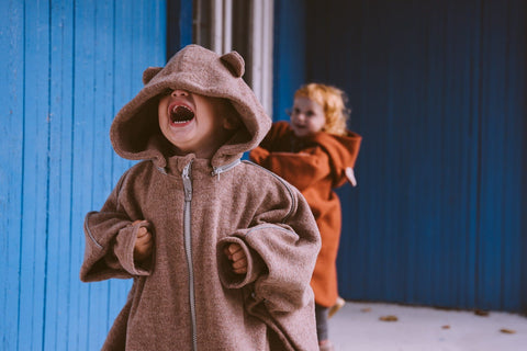 1. Two children wearing Patulove merino wool car ponchos with bear ears, playing outdoors against a blue wall