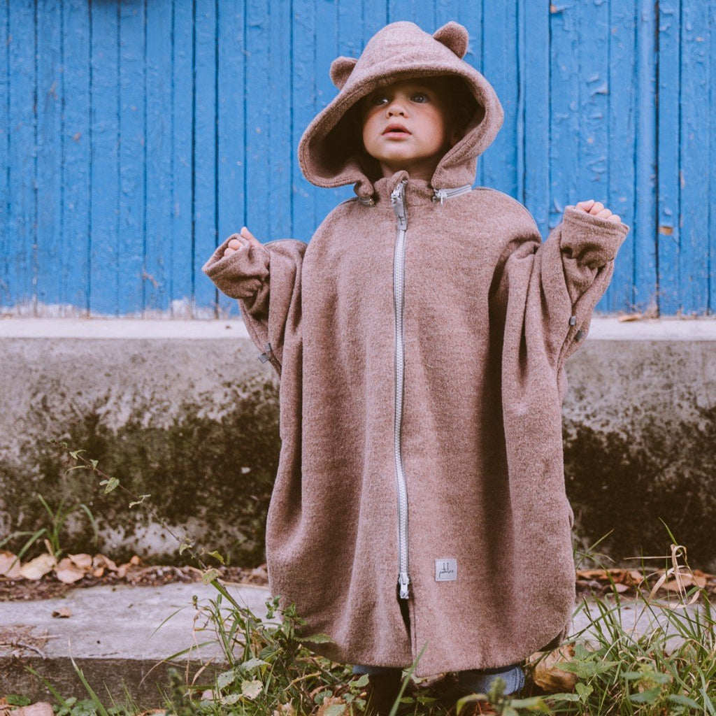 3. Child in brown Patulove merino wool car poncho with bear ears, standing outdoors with blue wall background