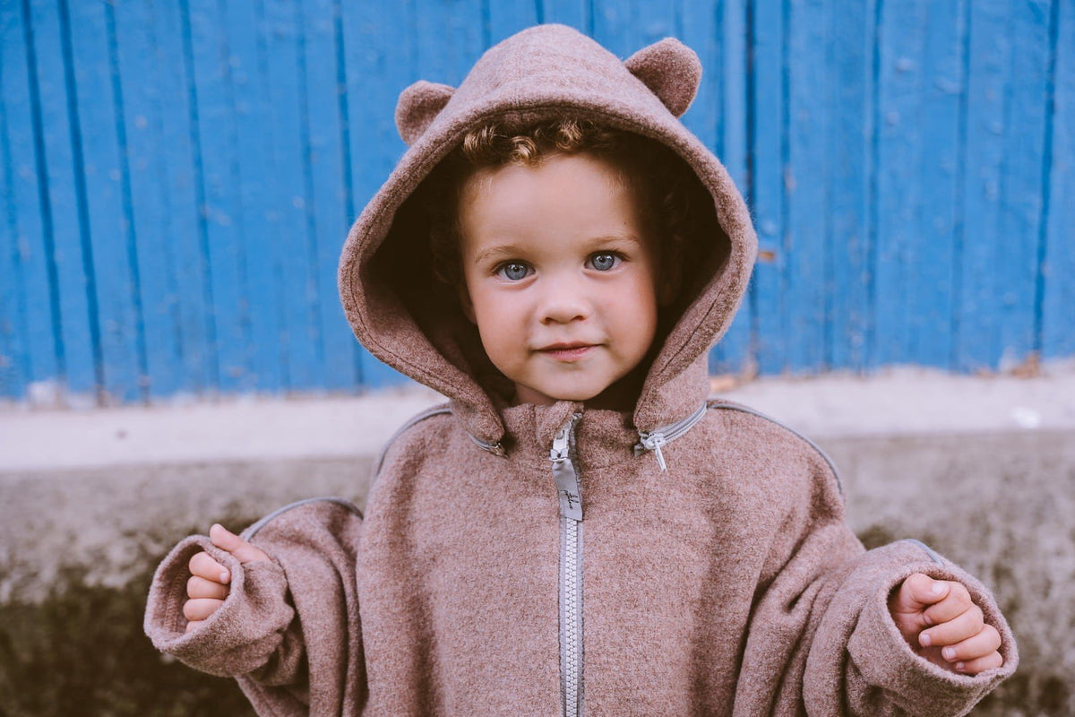 2. Child wearing brown Patulove merino wool car poncho with bear ears, standing in front of a blue wall