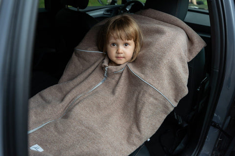 4. Child seated in car wearing brown Patulove merino wool car poncho, secured with car seat harness