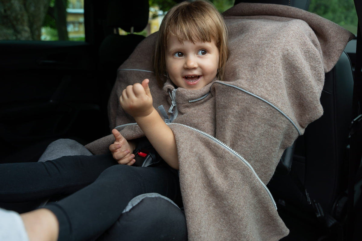 5. Child in car seat wearing brown Patulove merino wool car poncho, smiling and playing