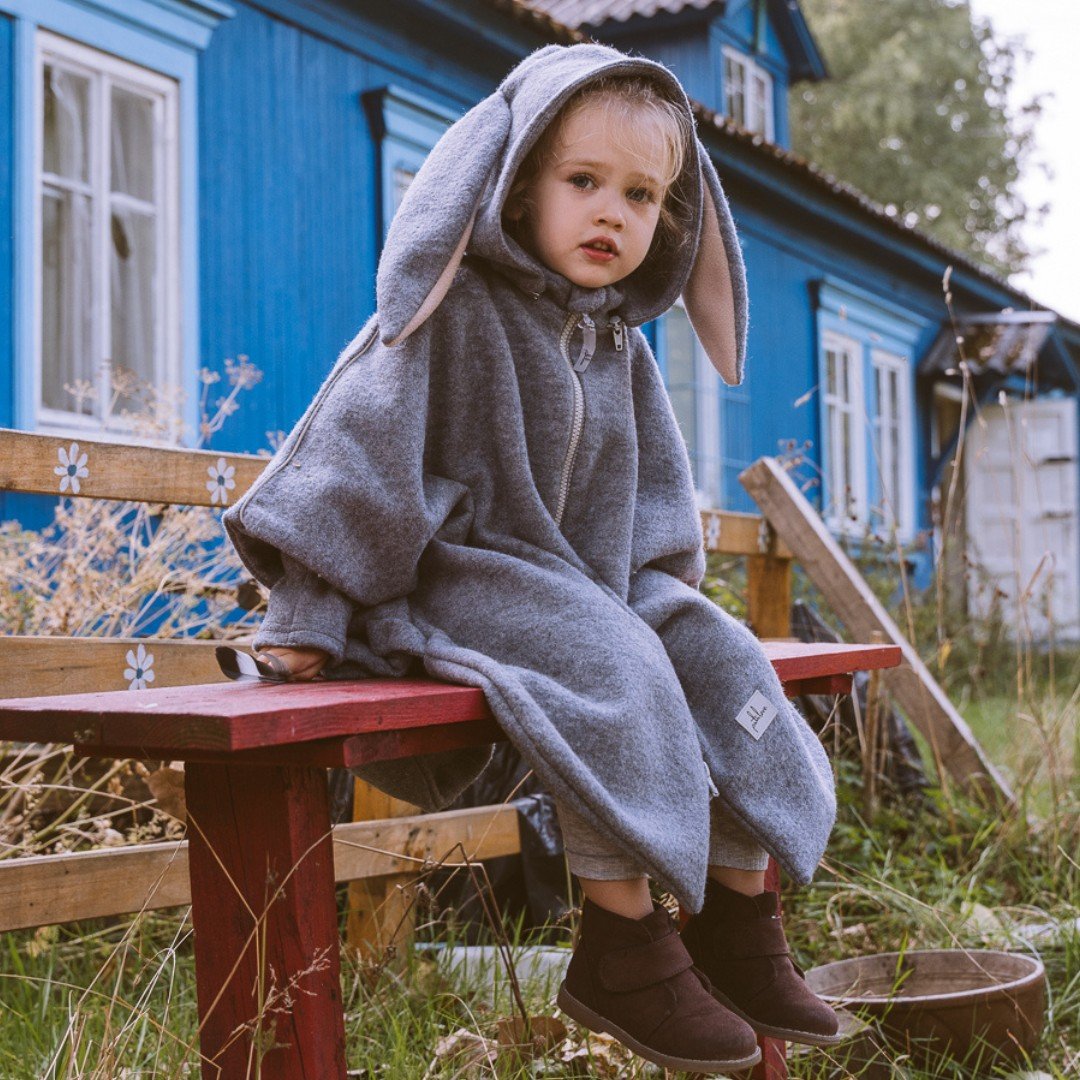 1. Child wearing grey merino wool poncho with bunny ears sitting on a bench in a rural setting