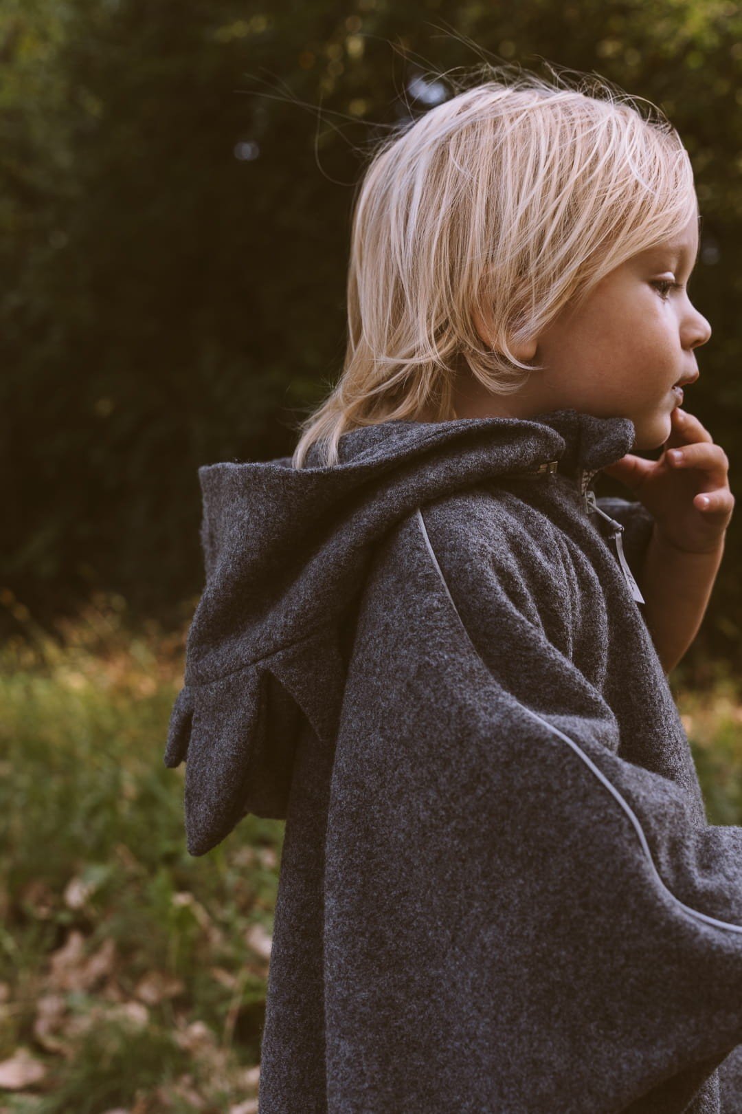 3. Side profile of child wearing grey merino wool poncho with dragon hood in a grassy area