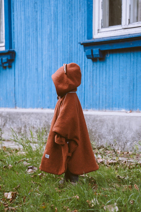 3. Side view of child in brown merino wool poncho with fox ears hood near blue house