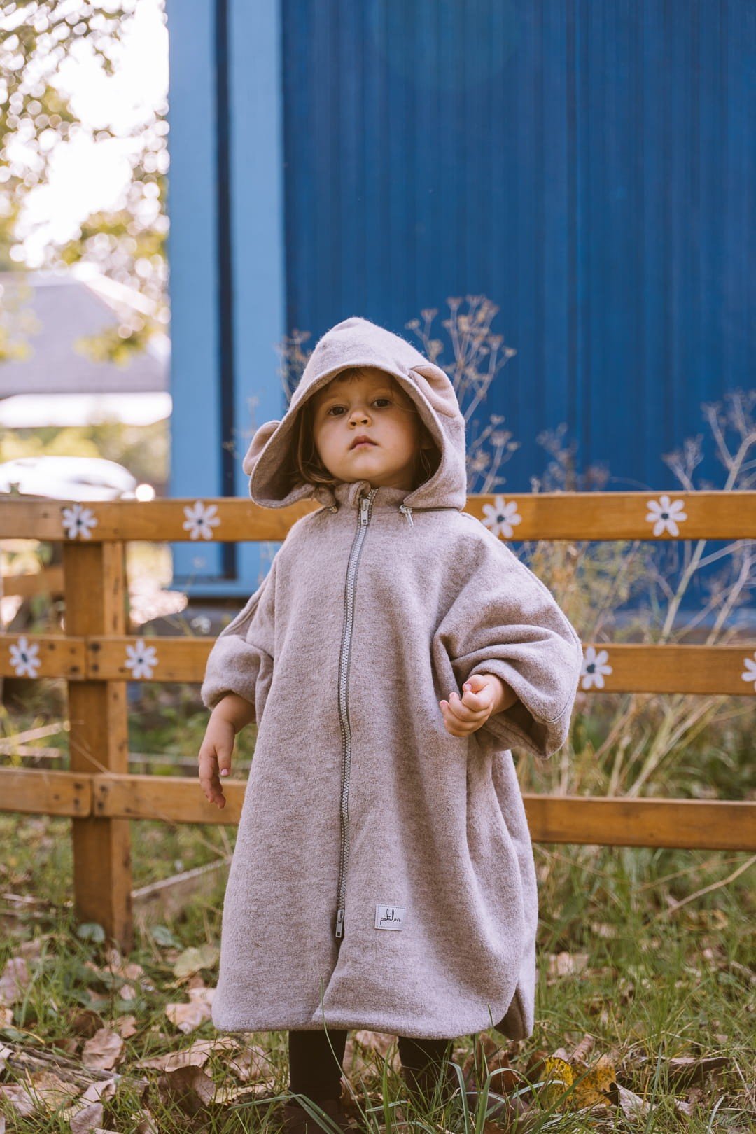 1. Child wearing beige merino wool poncho with hood in outdoor garden setting