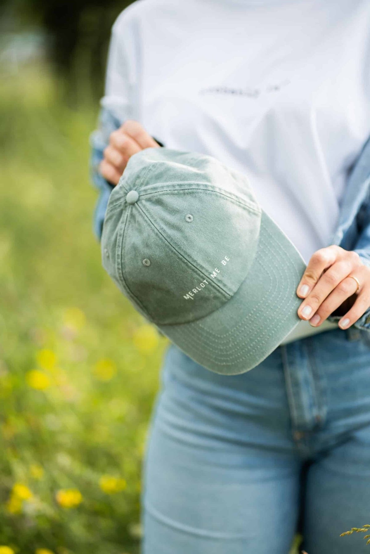 1. Woman holding green Merlot Me Be cap by Black Giraffe Brand in a grassy outdoor setting