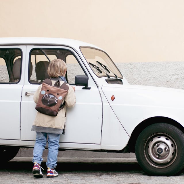 2. Child holding Muni bear backpack, wearing sunglasses and blue shirt, outdoor setting