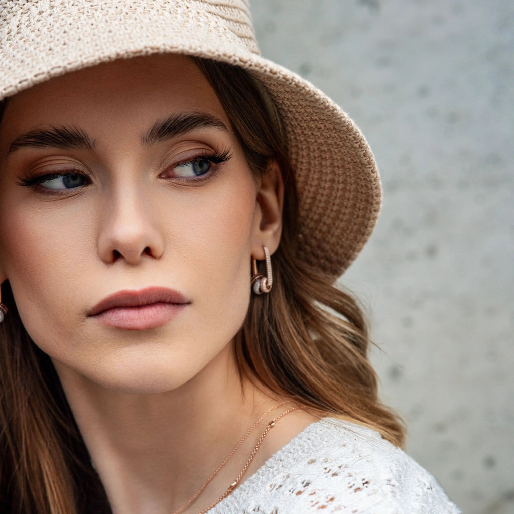 1. Woman wearing Metropolitan Glow white zircon charm earrings with rose gold plating, styled with a beige hat and white top, in an outdoor setting