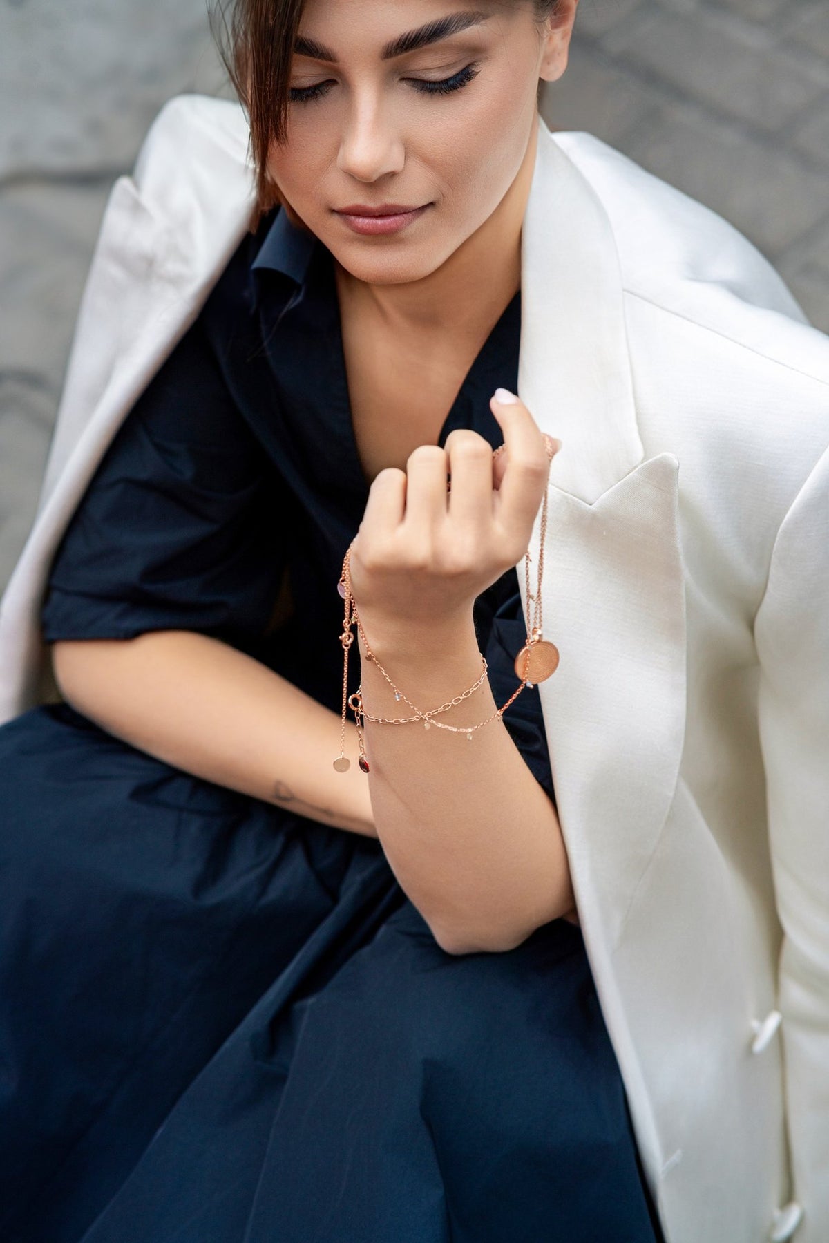 1. Woman in white blazer and navy dress holding rose gold Juno Mandala Necklace, sitting on urban pavement