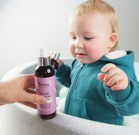 1. Baby in blue hoodie reaching for Nurme Mild Baby Body Lotion bottle held by an adult