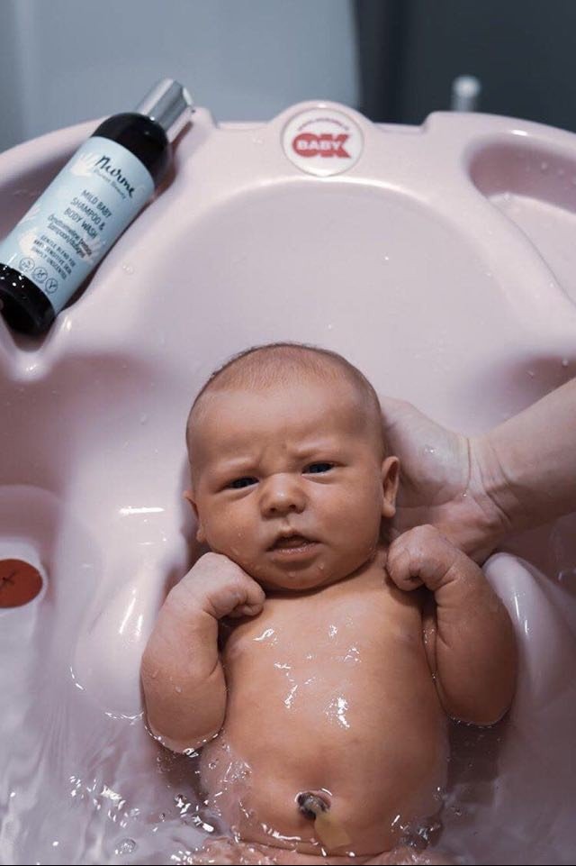 4. Baby in pink bath with Nurme Mild Baby Shampoo & Body Wash bottle on side, hand supporting baby