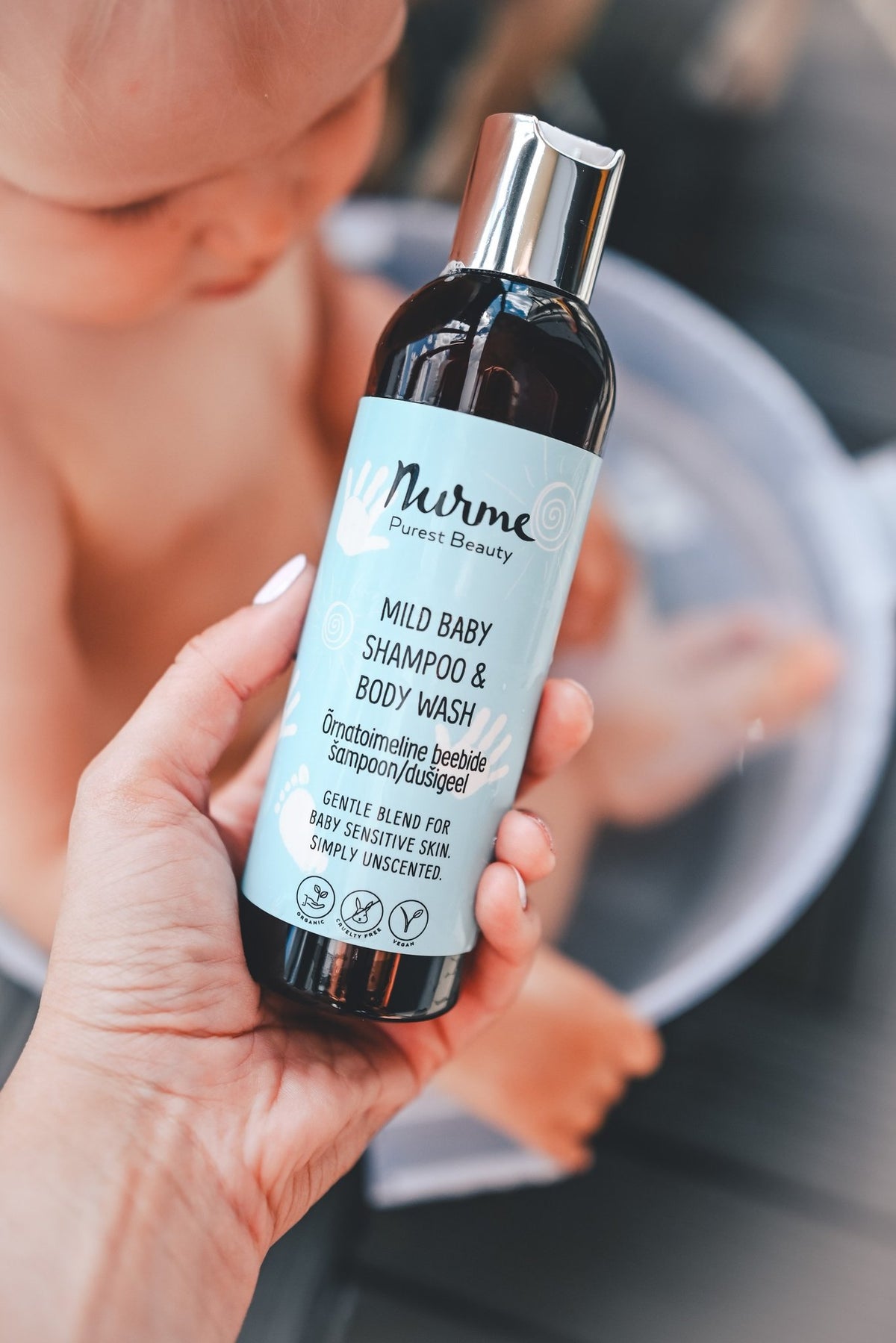 1. Hand holding Nurme Mild Baby Shampoo & Body Wash bottle with baby in background during bath time