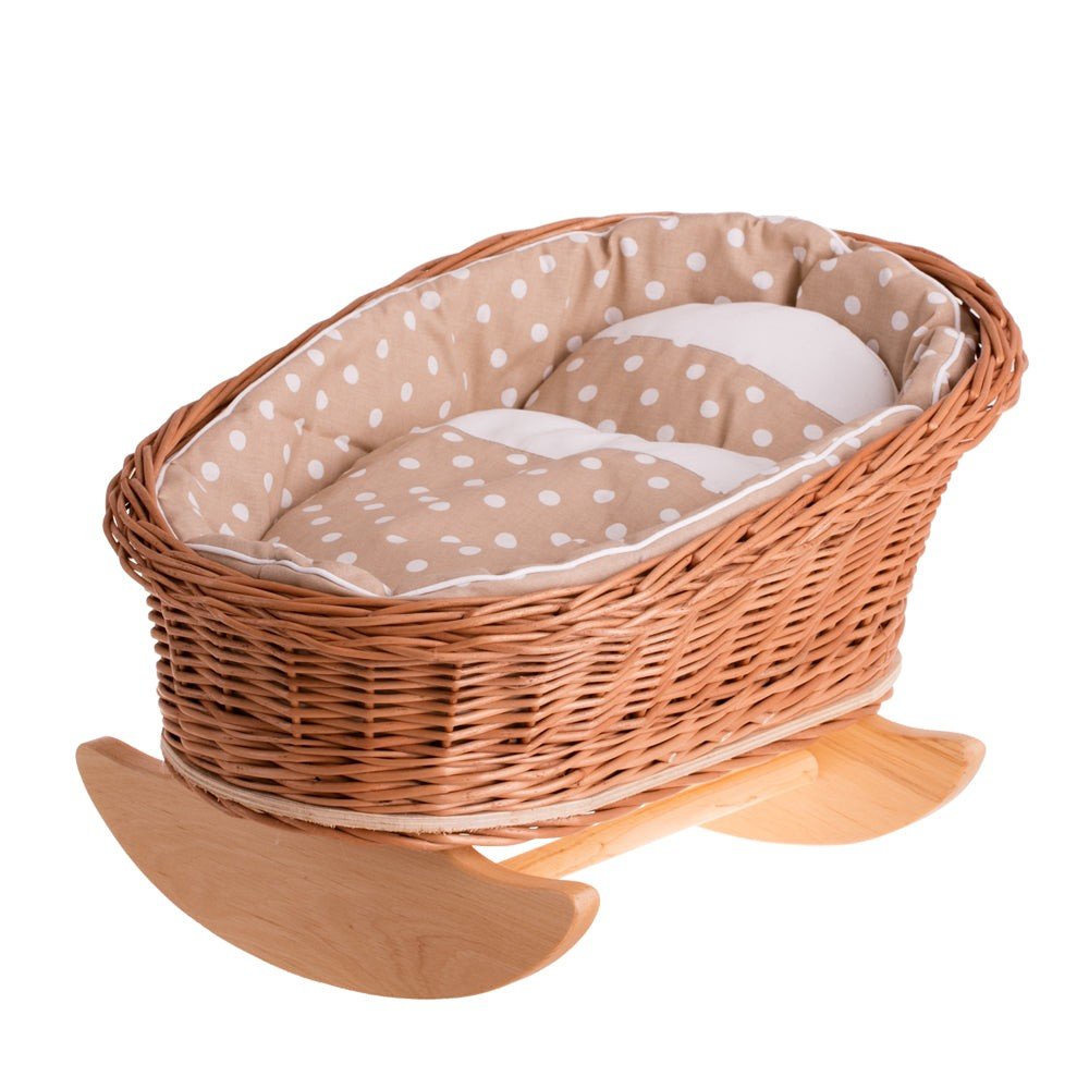 2. Wicker Stories doll cradle with beige polka dot lining, pillow, and quilt on wooden runners, showcasing traditional craftsmanship and natural materials in a studio setting.