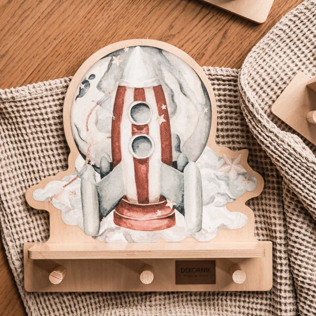 3. Close-up of rocketship mini wall shelf with colorful design and three wooden pegs, placed on a textured fabric surface.