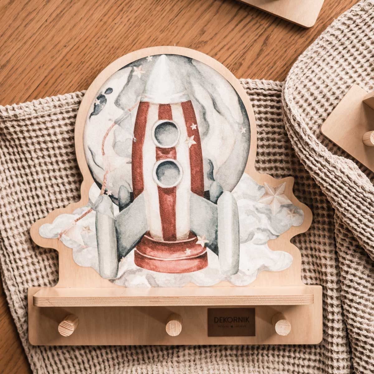 3. Close-up of rocketship mini wall shelf with colorful design and three wooden pegs, placed on a textured fabric surface.
