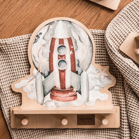 3. Close-up of rocketship mini wall shelf with colorful design and three wooden pegs, placed on a textured fabric surface.