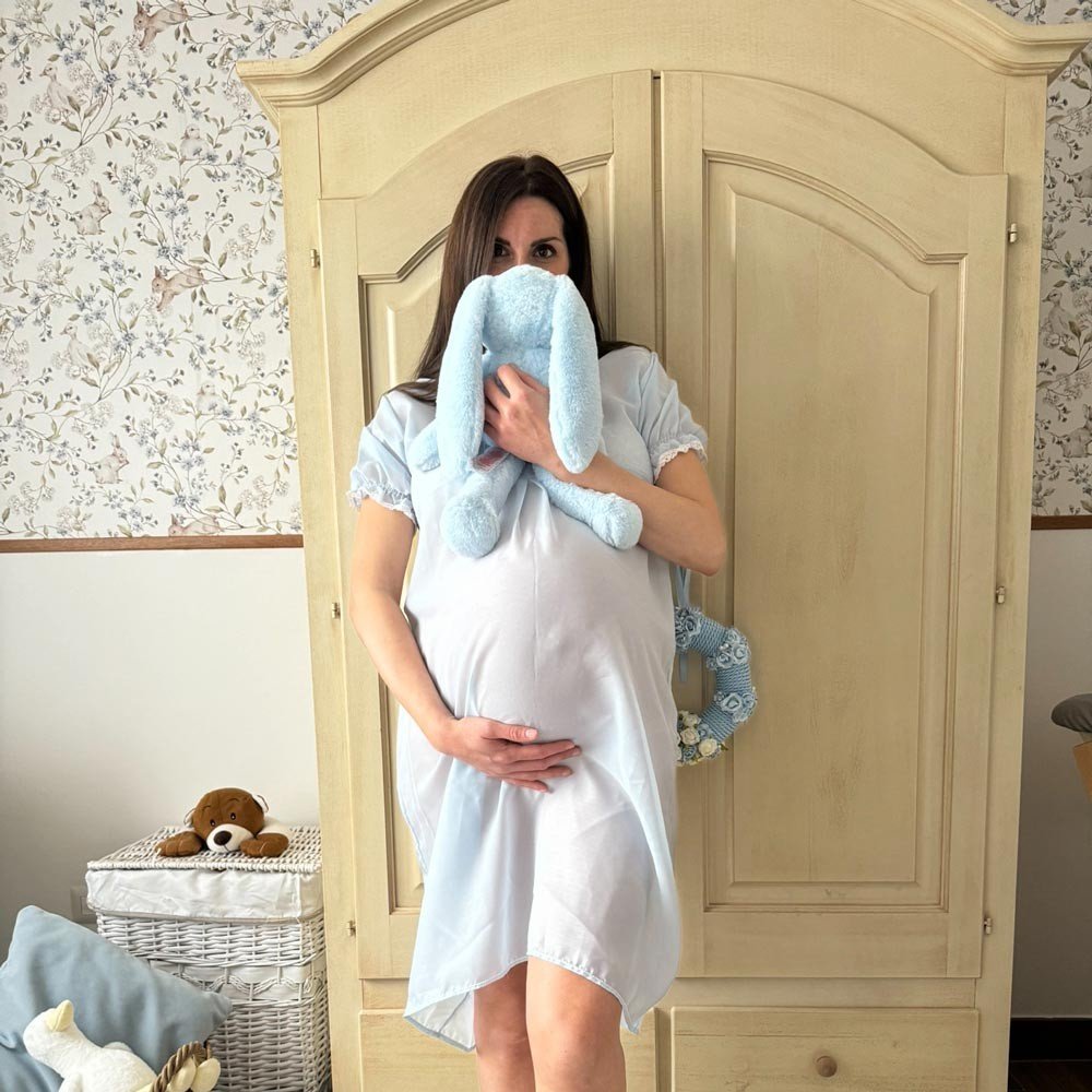 3. Pregnant woman in light blue dress holding plush bunny in front of beige wallpaper with ducks and bunnies