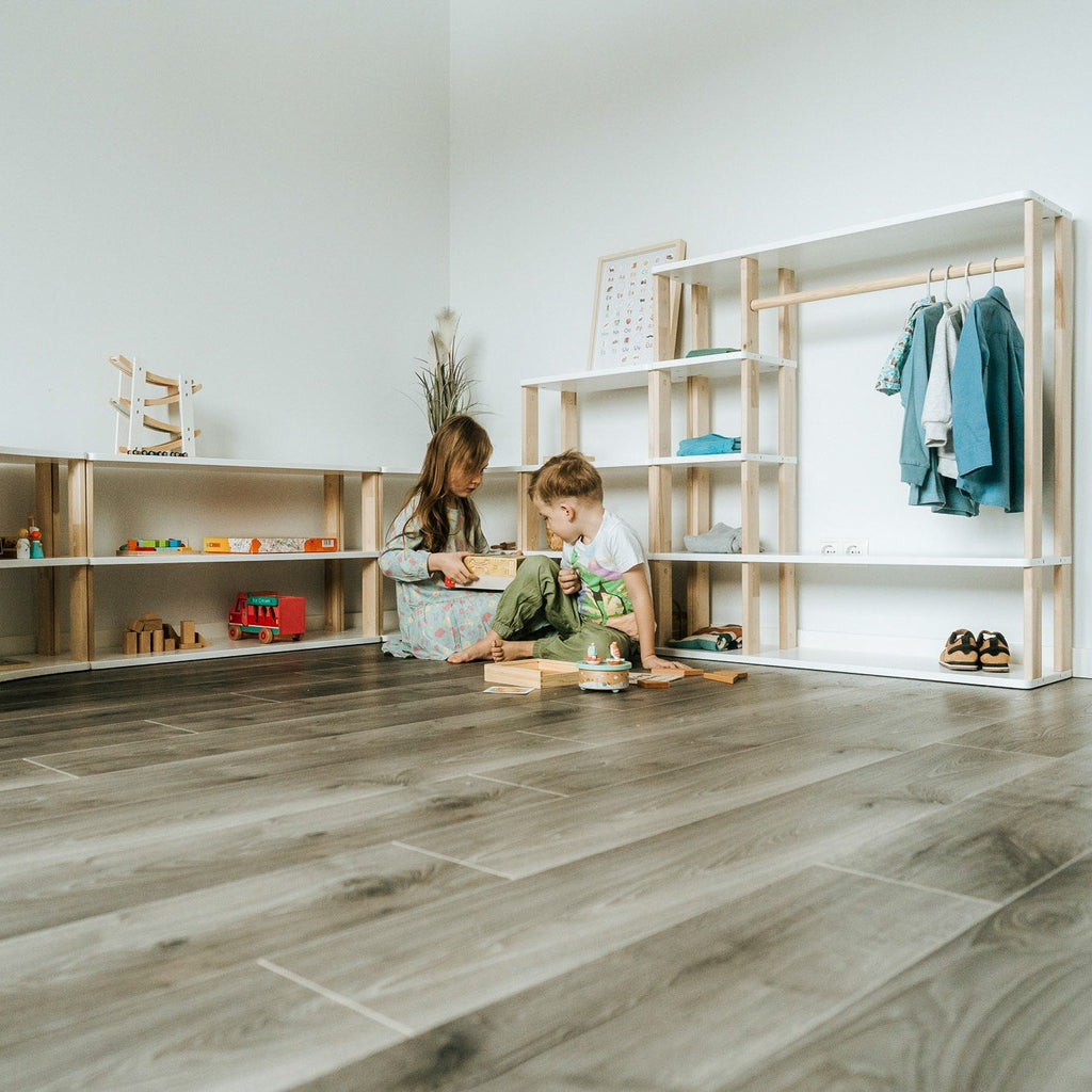 7. Two children playing near modular Montessori shelf with toys and clothes, set against a white wall on wooden flooring.