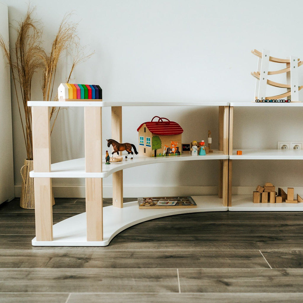 2. Corner view of modular Montessori shelf with three white shelves and natural wood supports, decorated with toys and books, on wooden flooring.