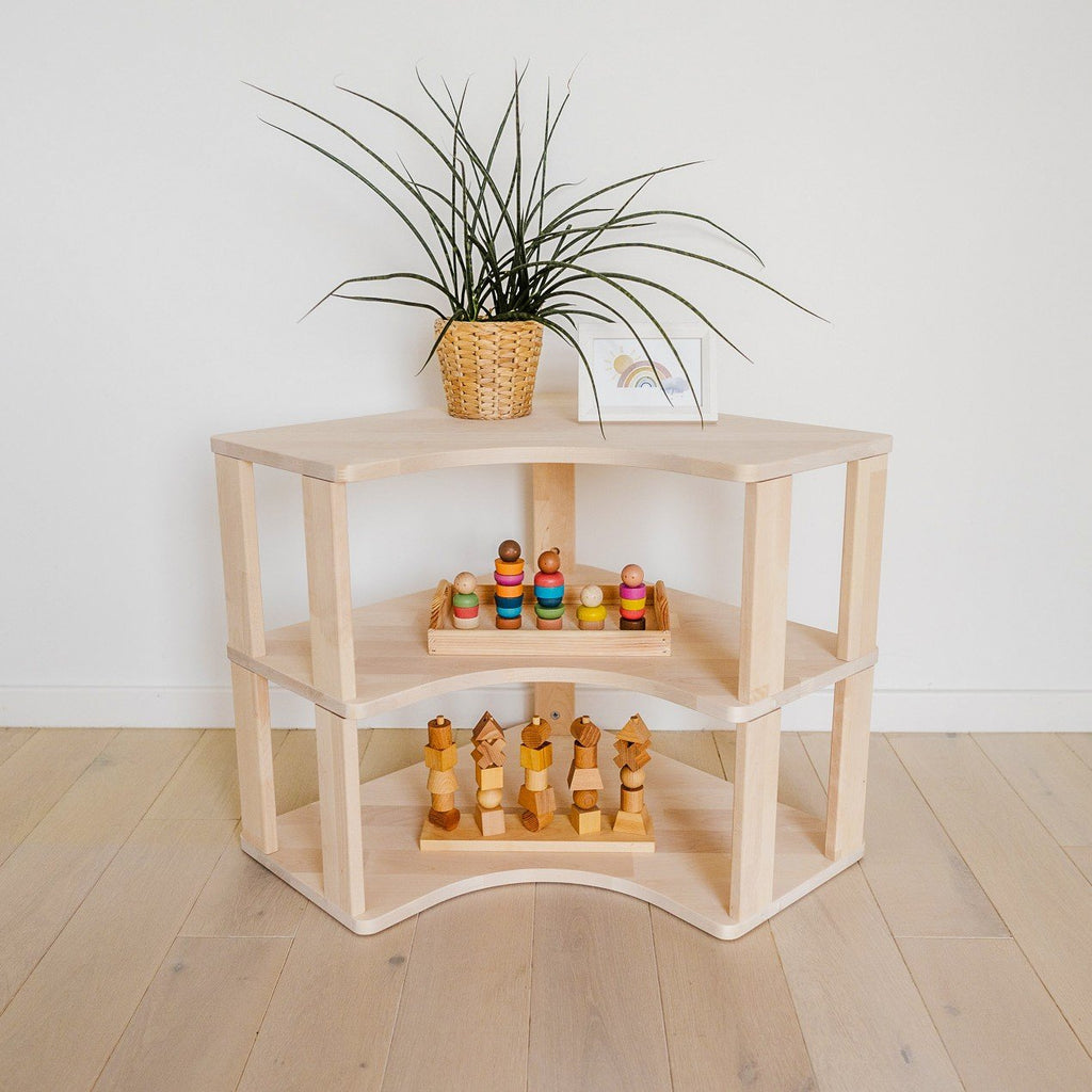 1. Natural wood Montessori corner shelf with two levels, featuring wooden toys and a potted plant on top, placed against a white wall on light wood flooring.