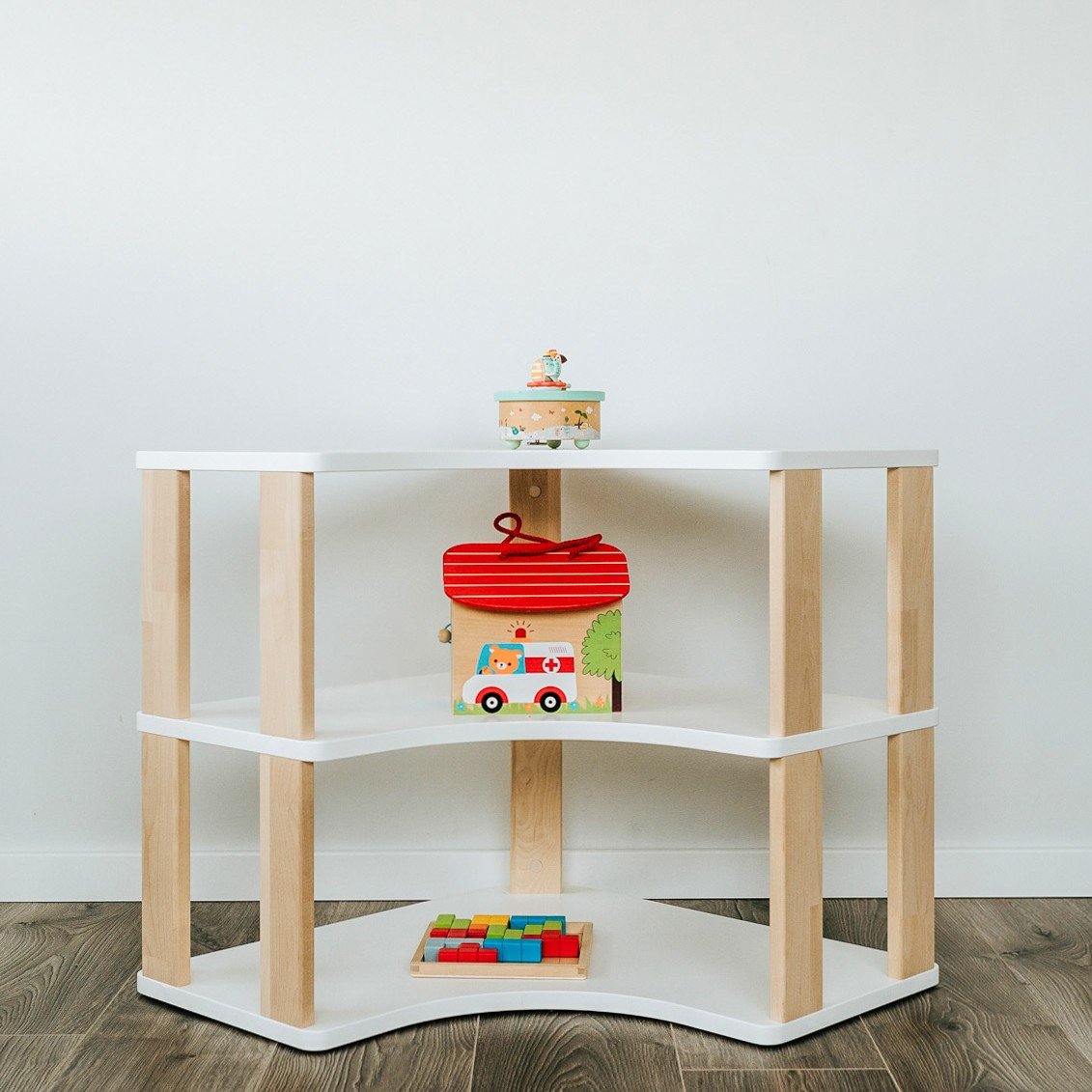 2. White and natural wood Montessori corner shelf with two levels, displaying colorful toys, set against a white wall on dark wood flooring.