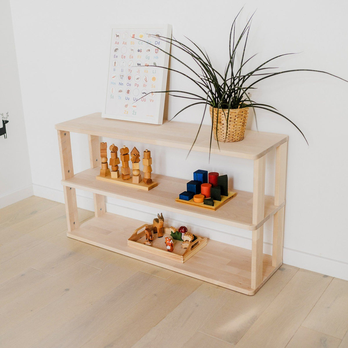 2. Three-tier Montessori shelf with wooden toys and a plant, placed in a minimalist room with light wood flooring