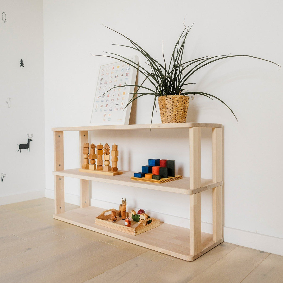 8. Three-shelf Montessori unit with toys and a plant, set against a decorated white wall with light wood flooring
