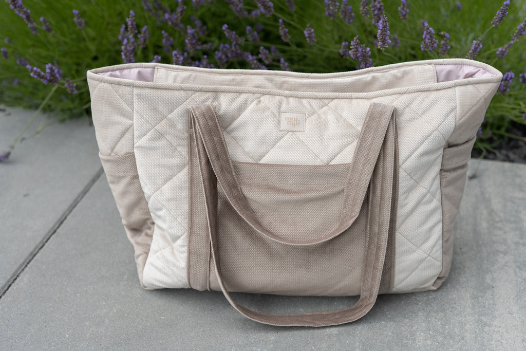 1. Beige mom weekender bag with quilted design and long straps on outdoor pavement with lavender plants