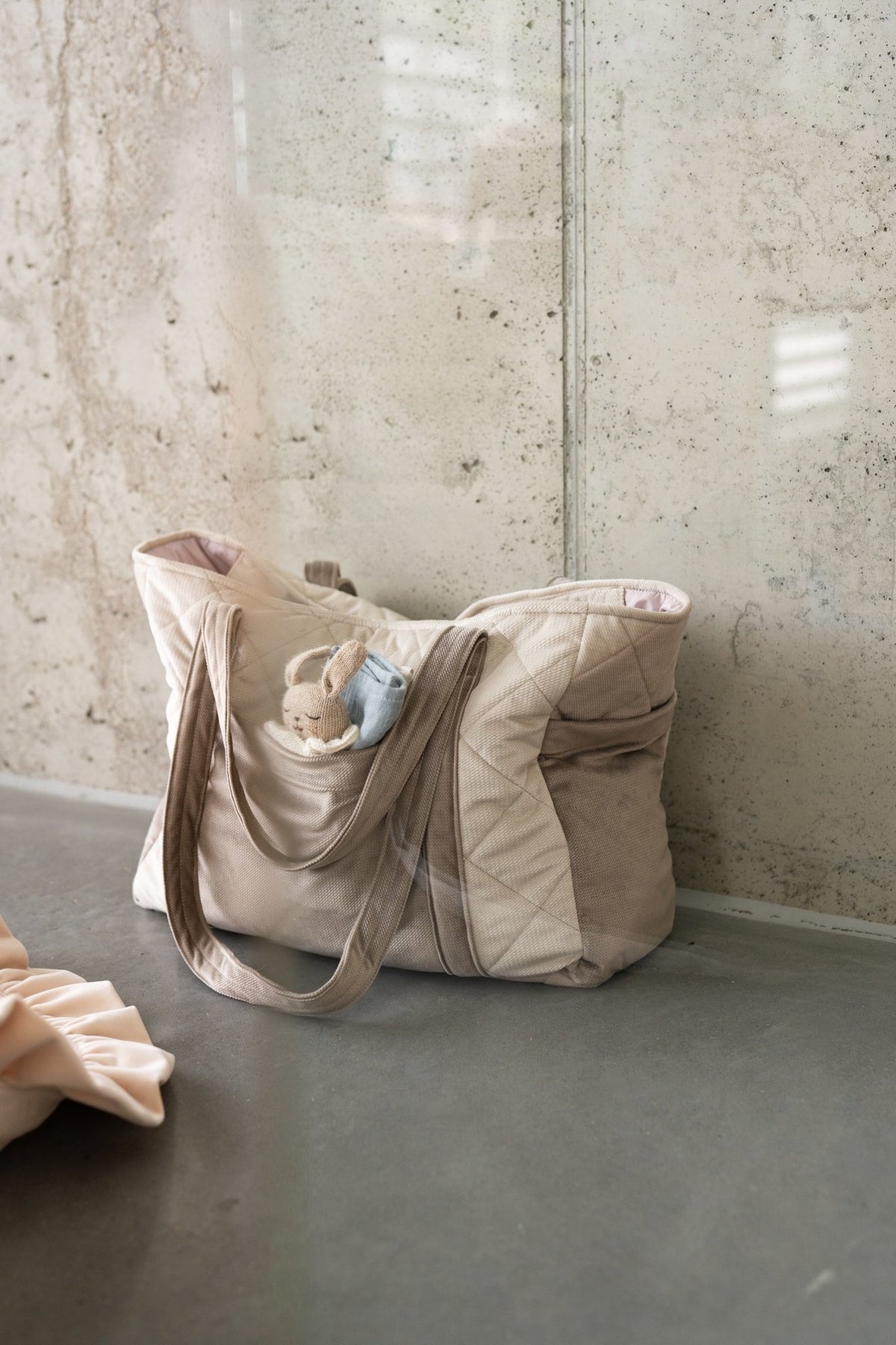 1. Beige mom weekender bag with long straps placed on concrete floor against a textured wall