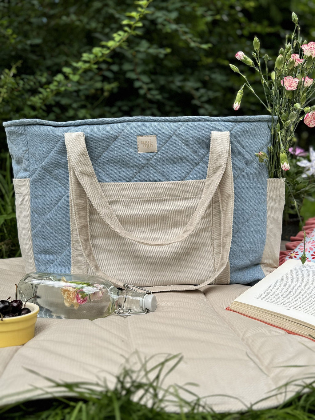 4. Blue and beige quilted weekender bag on a picnic blanket with flowers and a book in a garden setting