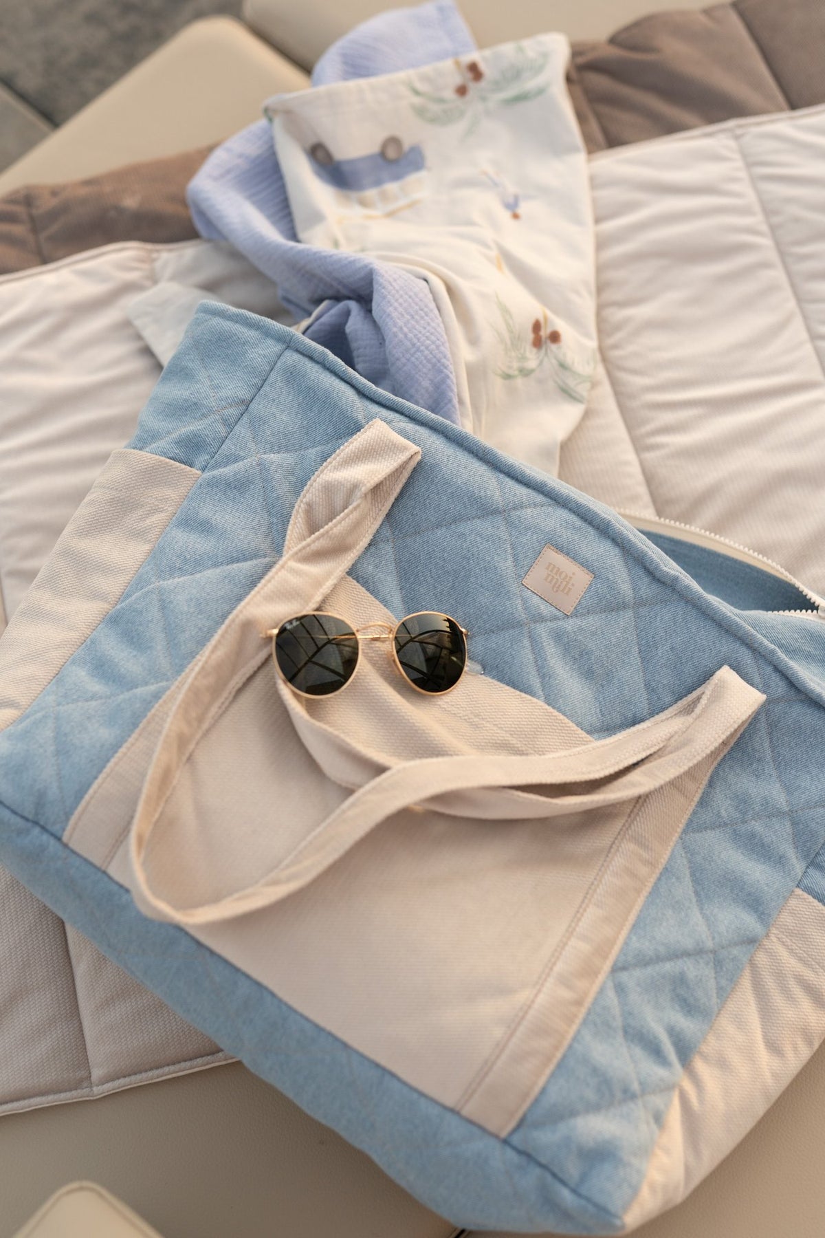 2. Lifestyle image of blue and beige weekender bag with sunglasses on top, placed on a beige outdoor cushion