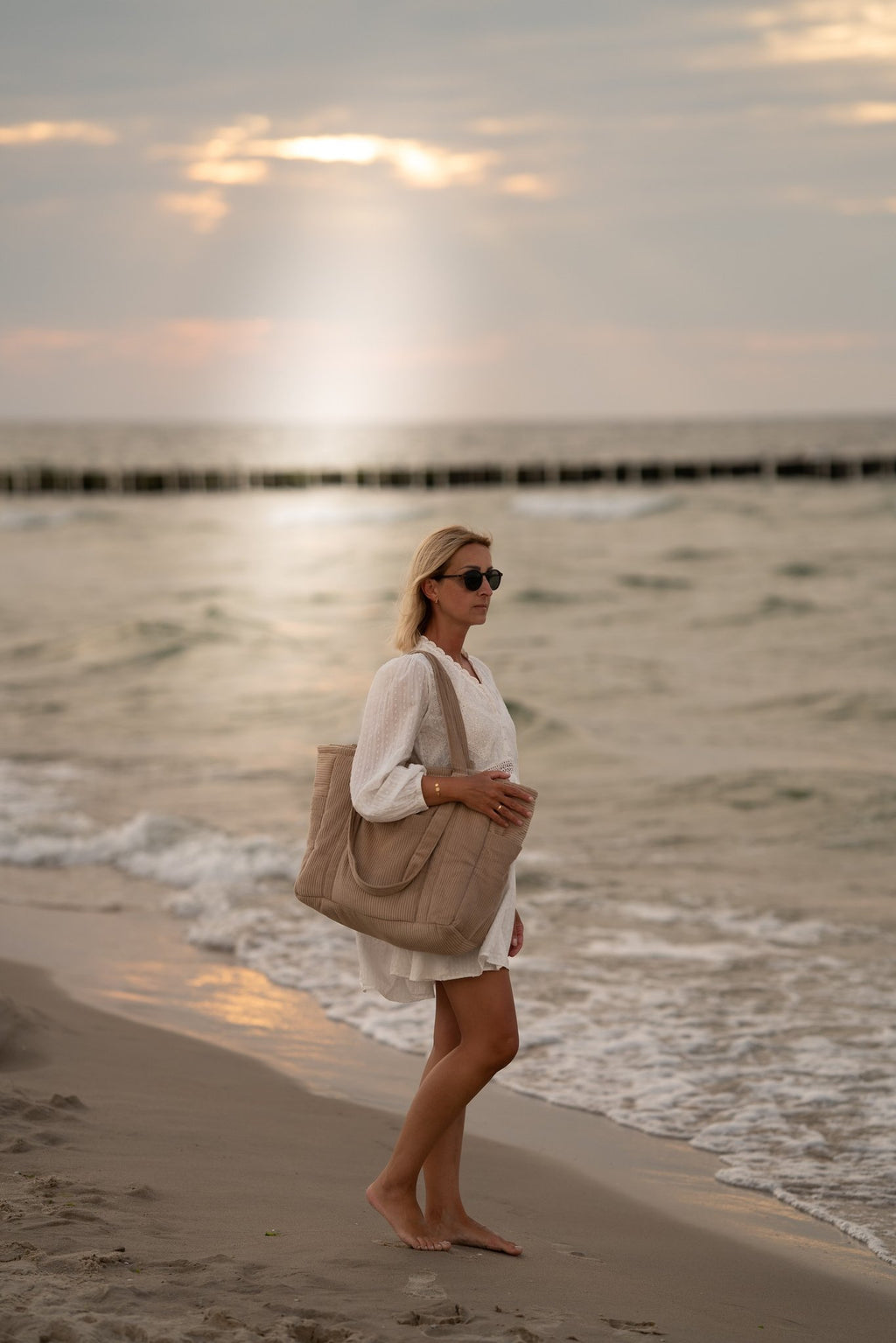 1. Woman walking on beach carrying latte-colored weekender bag, wearing white dress